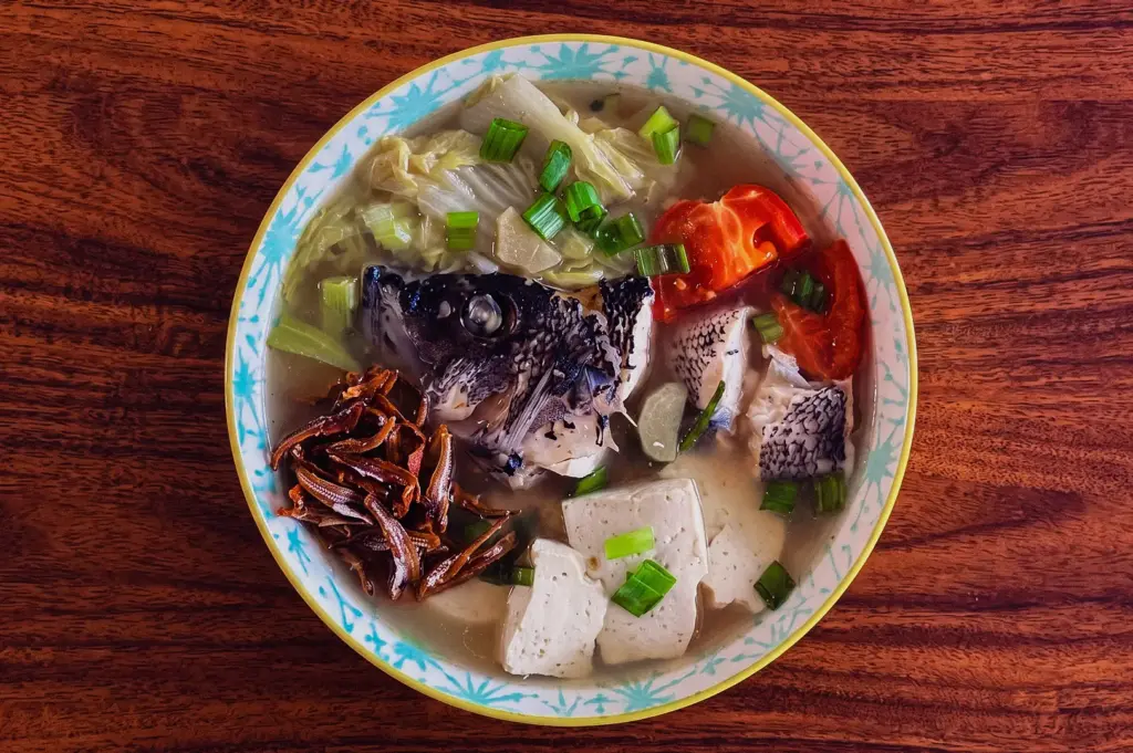 High-angle, close-up shot of a white spoon holding a tender slice of pomfret and julienned ginger over a steaming claypot of Hup Lok's Grandpa's Pomfret Fish Soup with tomatoes, shiitake mushrooms, and seaweed.