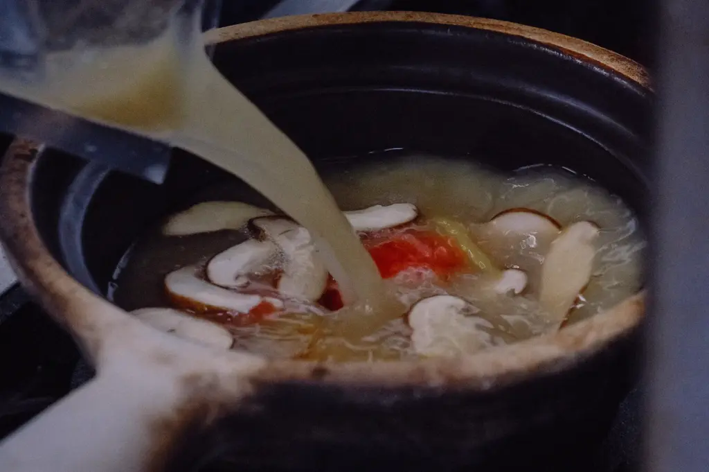 High-angle close-up of collagen-rich fish broth being poured into a claypot with pomfret fish soup ingredients, including shiitake mushrooms, ginger, and tomato, during preparation in a Singapore kitchen.