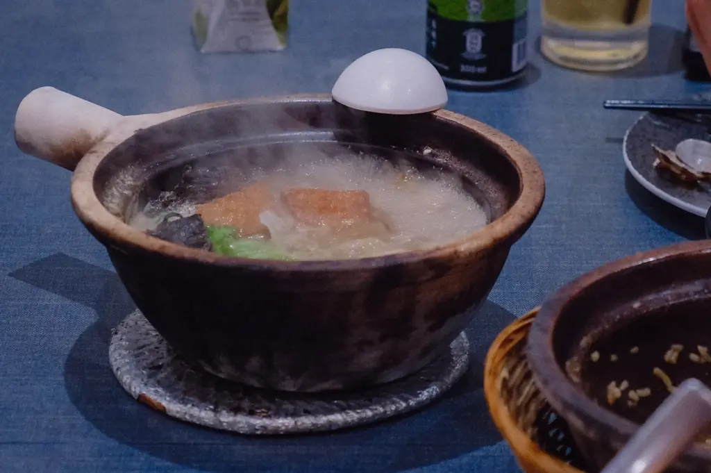 Wide eye-level table shot of a steaming claypot of pomfret fish soup resting on a trivet alongside side dishes and drinks.