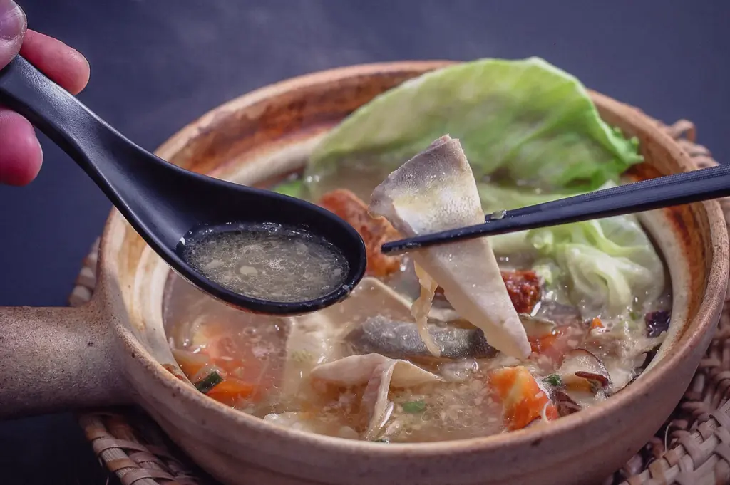 Eye-level close-up shot of tender pomfret fish being lifted from a claypot of Grandpa's Pomfret Fish Soup, featuring a clear collagen-rich broth with cabbage, tomato, shiitake mushrooms, and fresh ingredients at Hup Lok Singapore.
