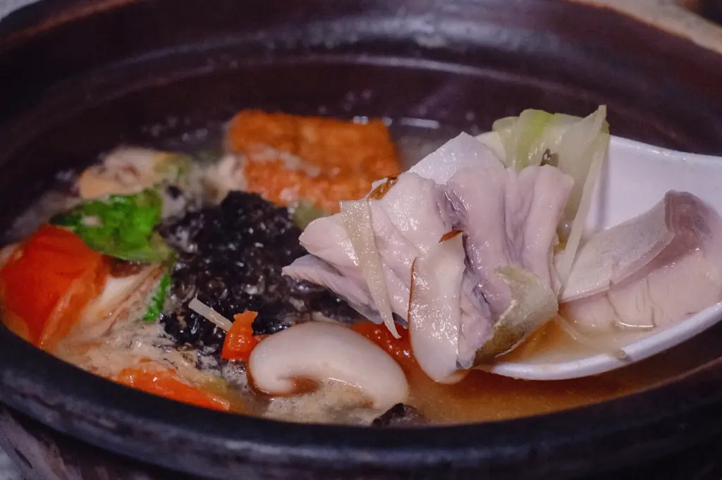 Close-up side angle of hearty fish soup in dark ceramic bowl, with rich collagen-thickened broth holding fish slices, mushrooms, seaweed, tomato, tofu, and leafy greens, as a white spoon lifts tender fish and vegetables above the surface, highlighting a robust pairing suited for rice or thick bee hoon that can carry the weight of the broth.