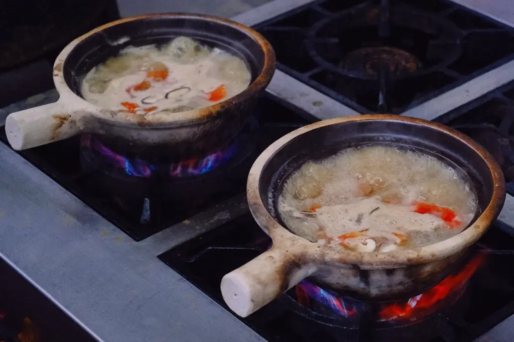 Traditional Singapore fish soup simmering in clay pots over open flames, featuring collagen-rich fish broth, fresh fish slices and vegetables, captured in a high-angle kitchen cooking shot.