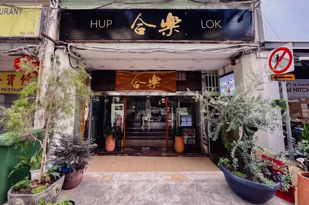 Straight-on, eye-level wide shot showcasing the traditional storefront exterior and carved wooden sign of the Hup Lok (合樂) fish soup restaurant along Havelock Road in Singapore.