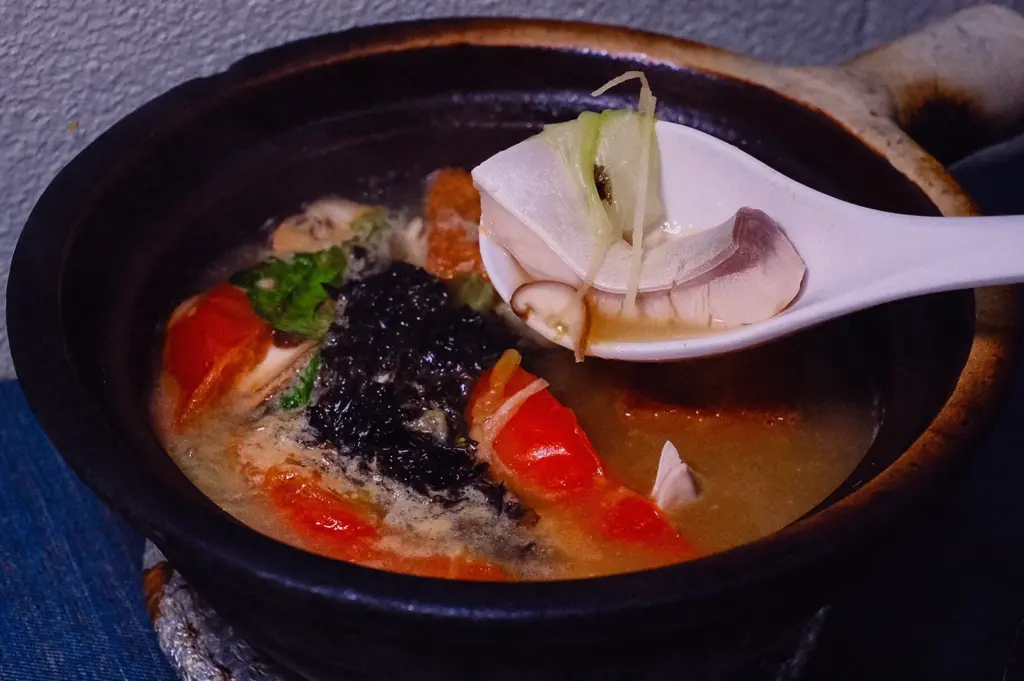 High-angle, close-up shot of a white spoon holding a tender slice of pomfret and julienned ginger over a steaming claypot of Hup Lok's Grandpa's Pomfret Fish Soup with tomatoes, shiitake mushrooms, and seaweed.
