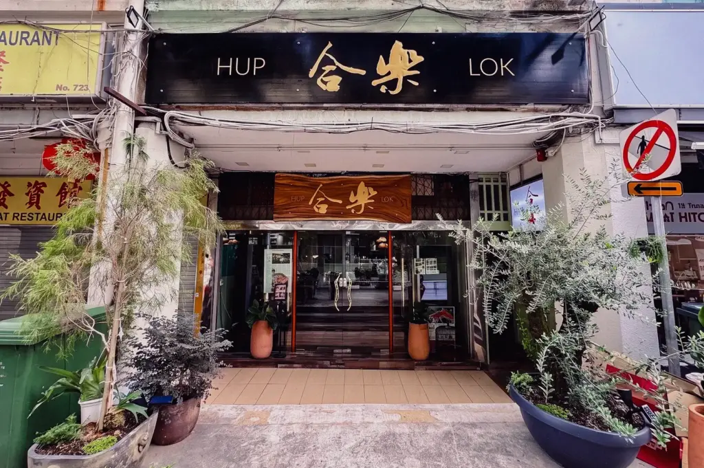 Straight-on, eye-level wide shot showcasing the traditional storefront exterior and carved wooden sign of the Hup Lok (合樂) fish soup restaurant along Havelock Road in Singapore.