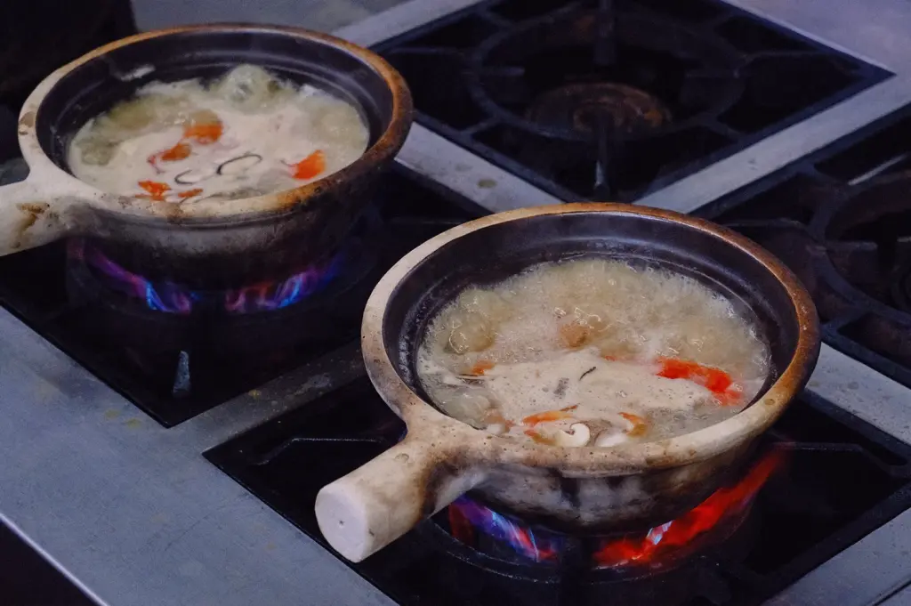 Three-quarter angle view of collagen-rich fish soup broth simmering in clay pots over open flames, showcasing traditional fish bone stock preparation for Singapore fish soup.