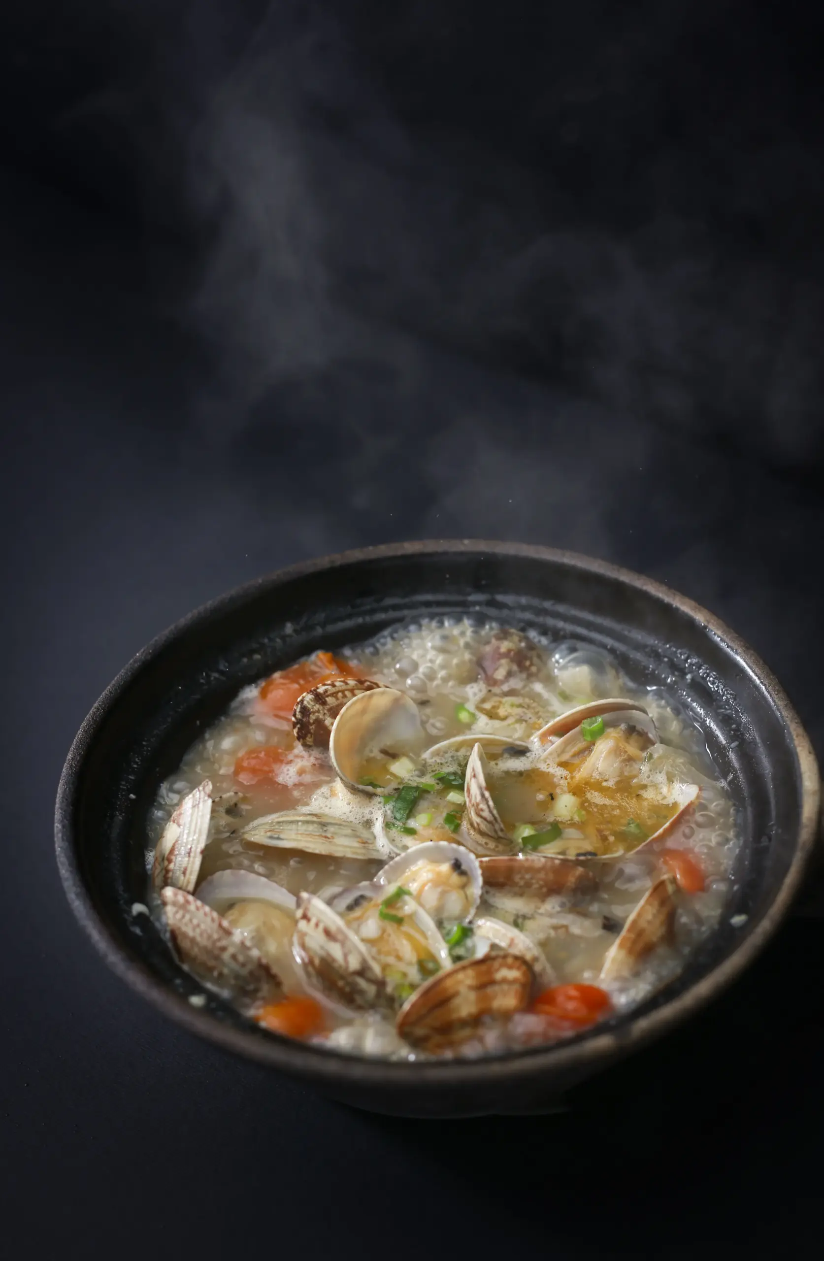 Close-up overhead angle of steaming clams in white wine broth served in a rustic black clay pot, garnished with chopped spring onions and tomatoes, showcasing a fresh seafood soup with rich savory flavors.