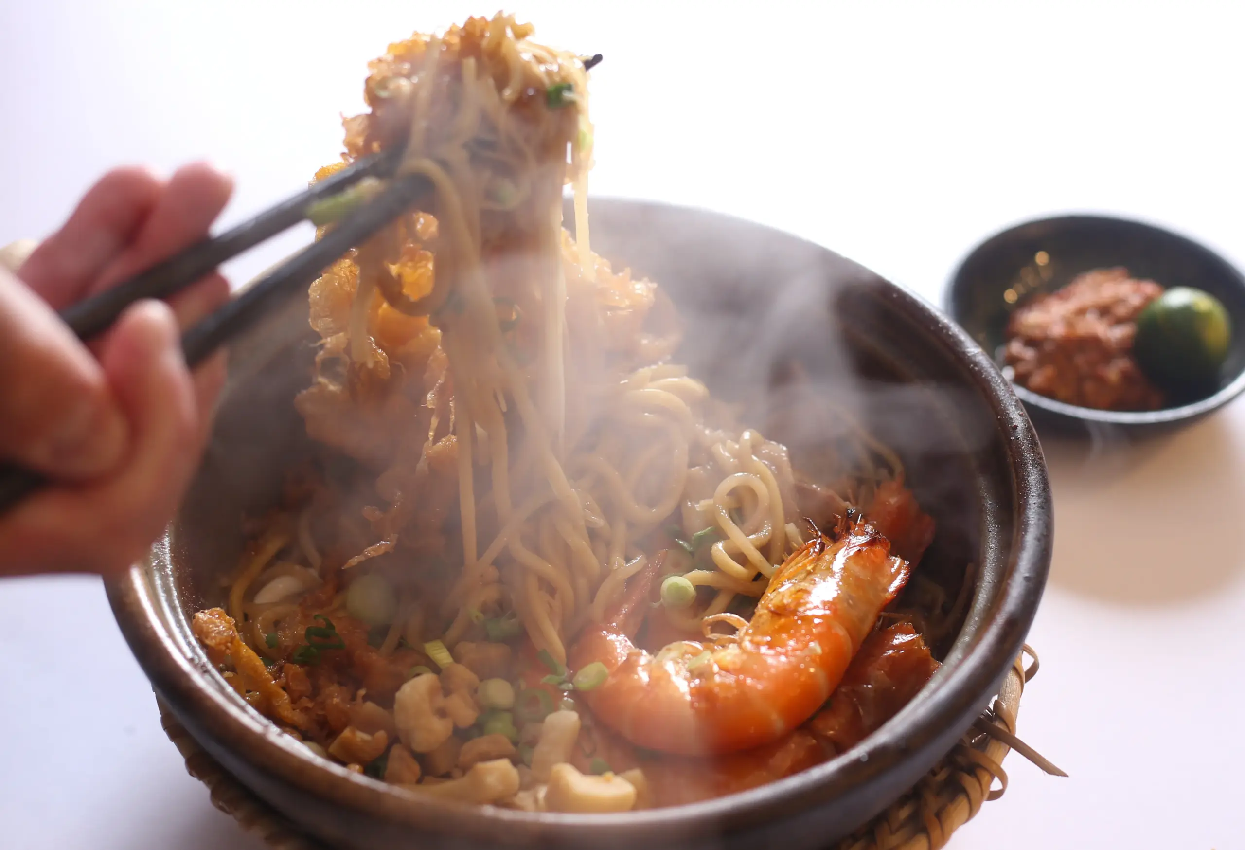 Close-up three-quarter angle shot of claypot Hokkien mee with noodles lifted by chopsticks, featuring prawns, vegetables, and rising steam in a traditional clay pot for an authentic Malaysian seafood noodle experience.