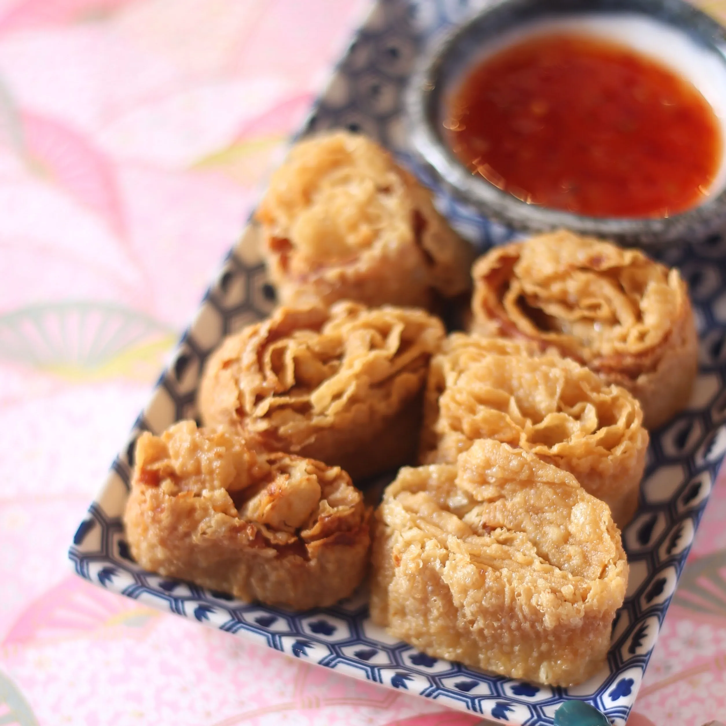 Top‑down shot of a rectangular white ceramic plate holding six golden brown fried bean curd rolls, also known as tofu skin rolls or yuba rolls, arranged neatly beside a small black dish of dark soy‑based dipping sauce, set against a gray tabletop background, highlighting the crispy layered texture and authentic Asian dim sum appetizer presentation.