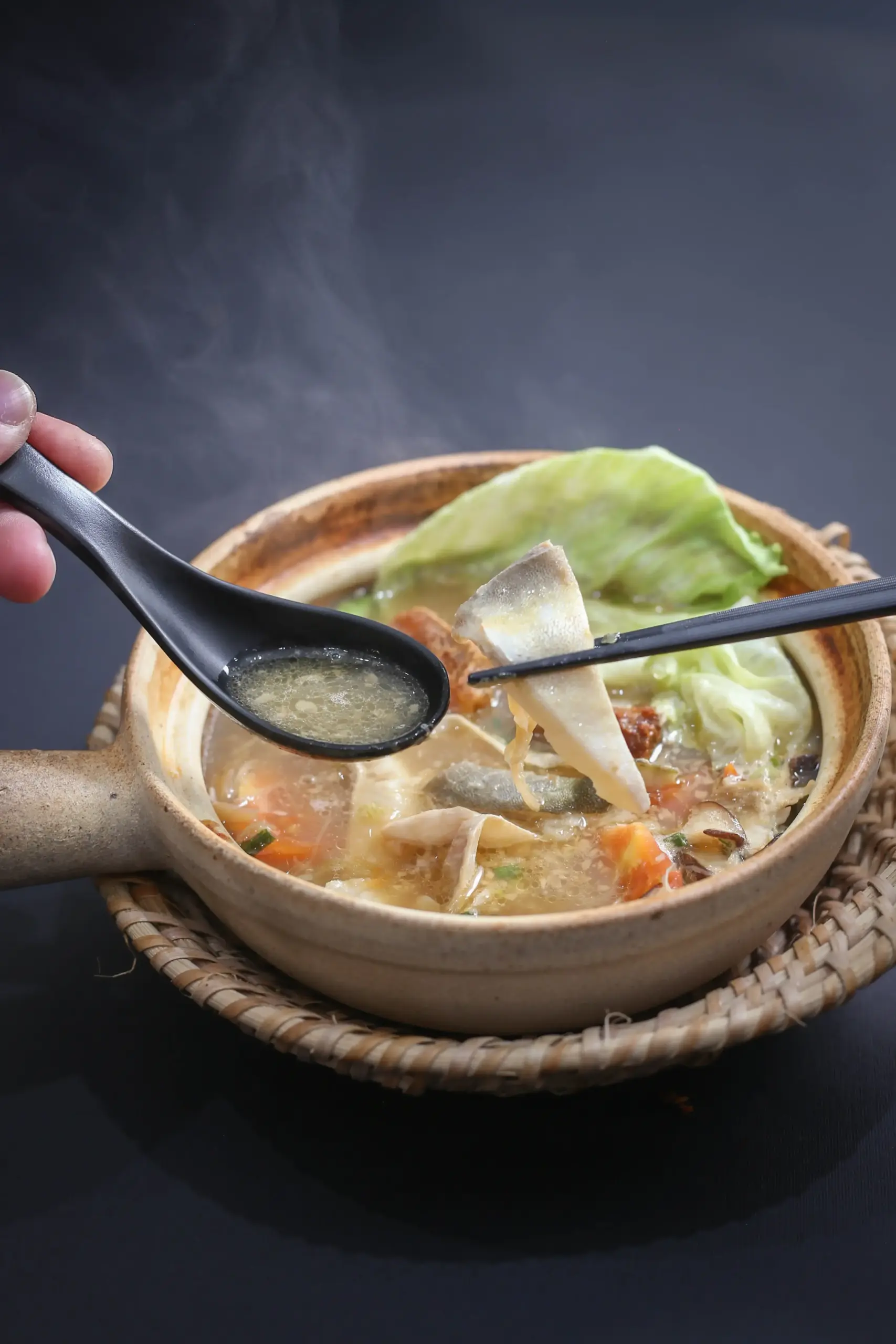 Close-up high-angle shot of pomfret fish soup in a traditional clay pot, with fish slices held by chopsticks above a light broth, accompanied by fresh vegetables and a soup spoon highlighting a healthy Asian seafood dish.