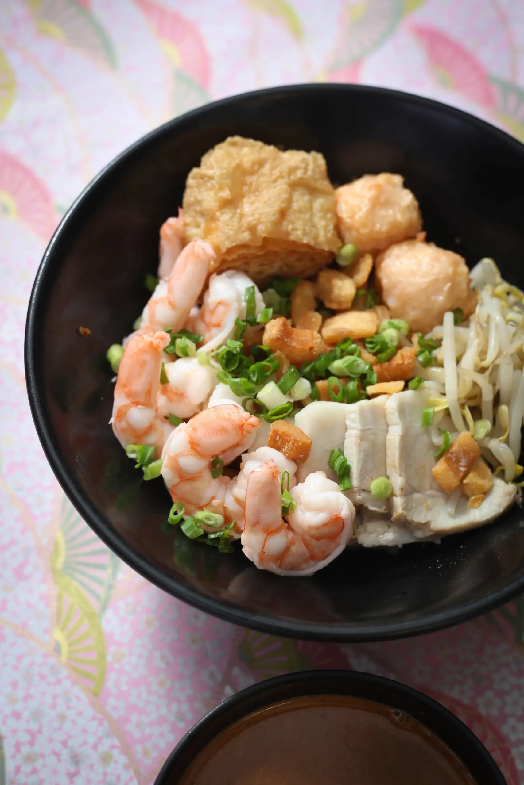 Top‑down shot of a black bowl filled with Asian noodle soup ingredients, including cooked shrimp, tofu cubes, fried tofu, fish balls, bean sprouts, chopped scallions, and crispy fried garlic, placed on a pastel patterned surface with fan motifs, highlighting vibrant textures and authentic Asian street‑food style.