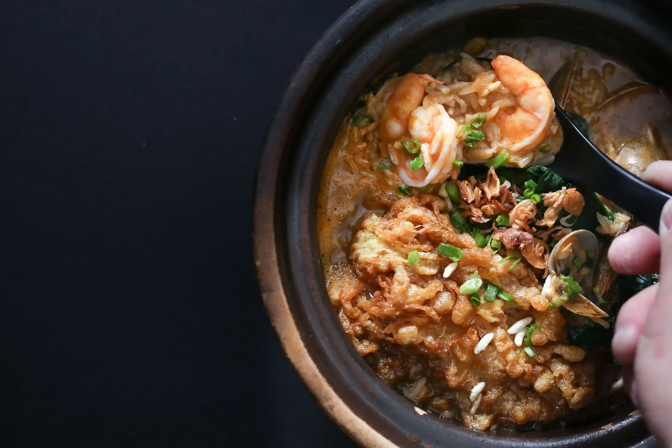 Three-quarter overhead close-up shot of seafood pao fan in a clay pot, with a spoon lifting fresh prawns above crispy rice, clams, spring onions, and rich seafood broth, highlighting a comforting Cantonese-style rice soup.