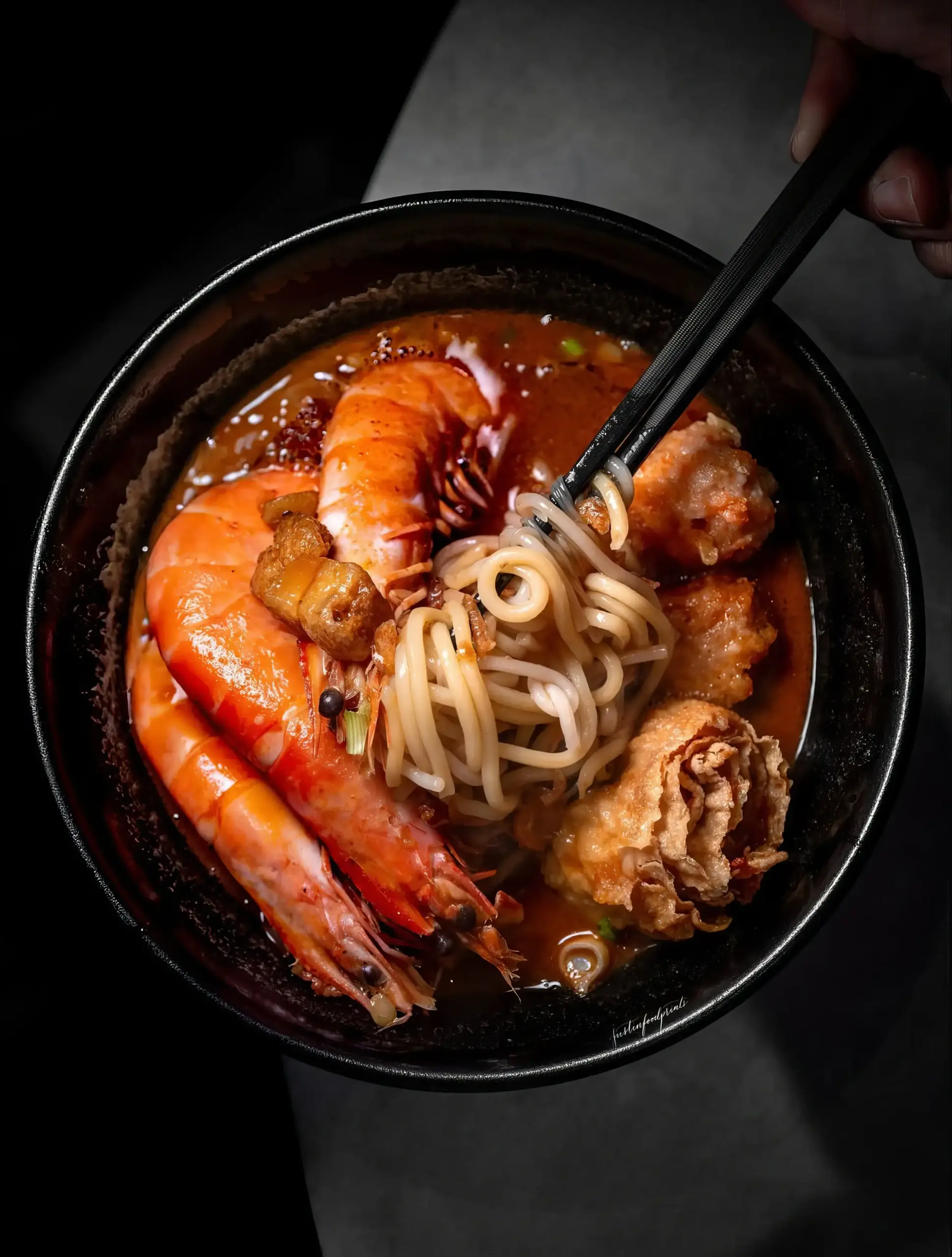 Close‑up angled shot of a steaming bowl of reddish‑brown seafood broth with noodles lifted by chopsticks, large prawns, and fried bean curd skin, showcasing rich colors and textures of Southeast Asian prawn noodle soup in an appetizing presentation.