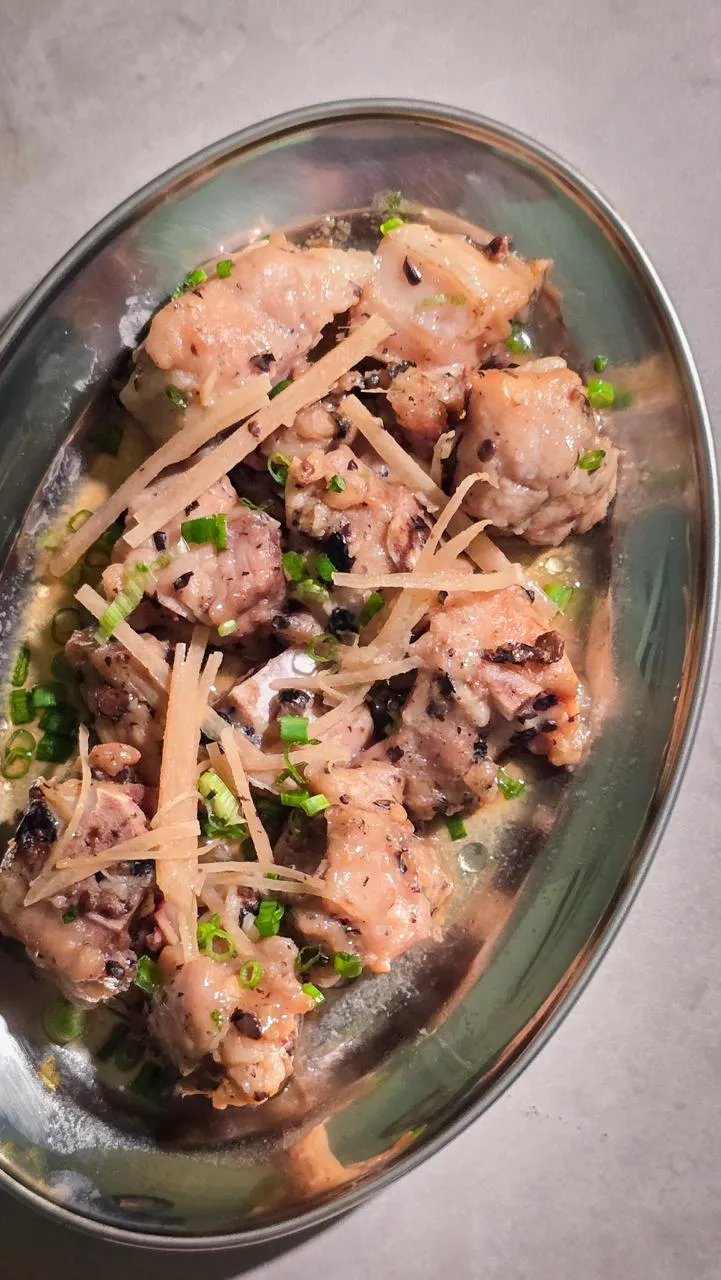 Close-up overhead 45-degree angle shot of traditional Cantonese steamed pork ribs (蒸排骨), garnished with fresh ginger strips and chopped scallions, served in a stainless steel dish with savory sauce at a dim sum restaurant.