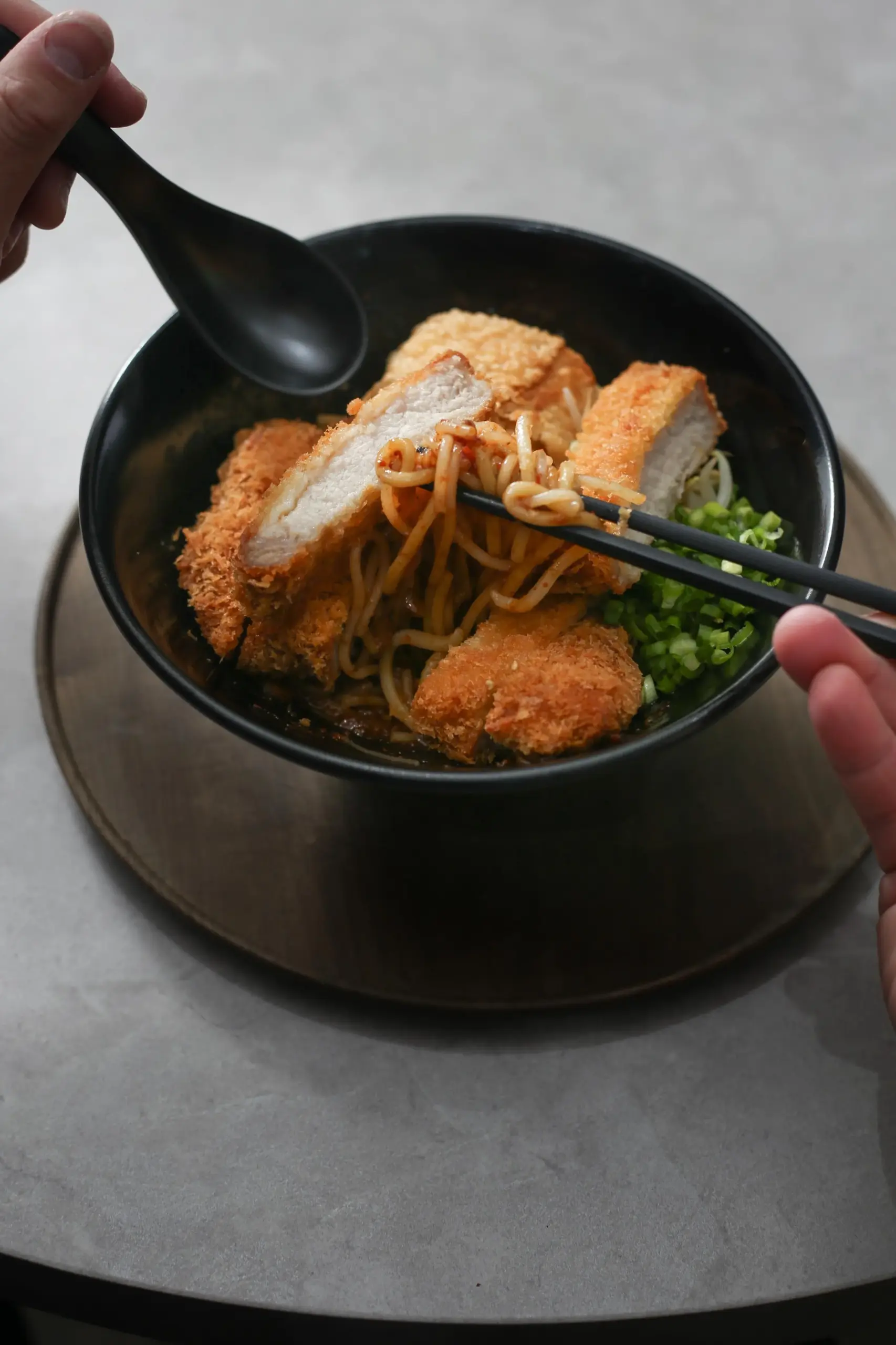 Close‑up angled shot of Japanese ramen noodles lifted with black chopsticks, topped with golden breaded chicken katsu cutlets, chopped green onions, and bean sprouts in a savory broth, highlighting the glossy noodles and crispy texture of the fried chicken in a comforting Japanese dish.