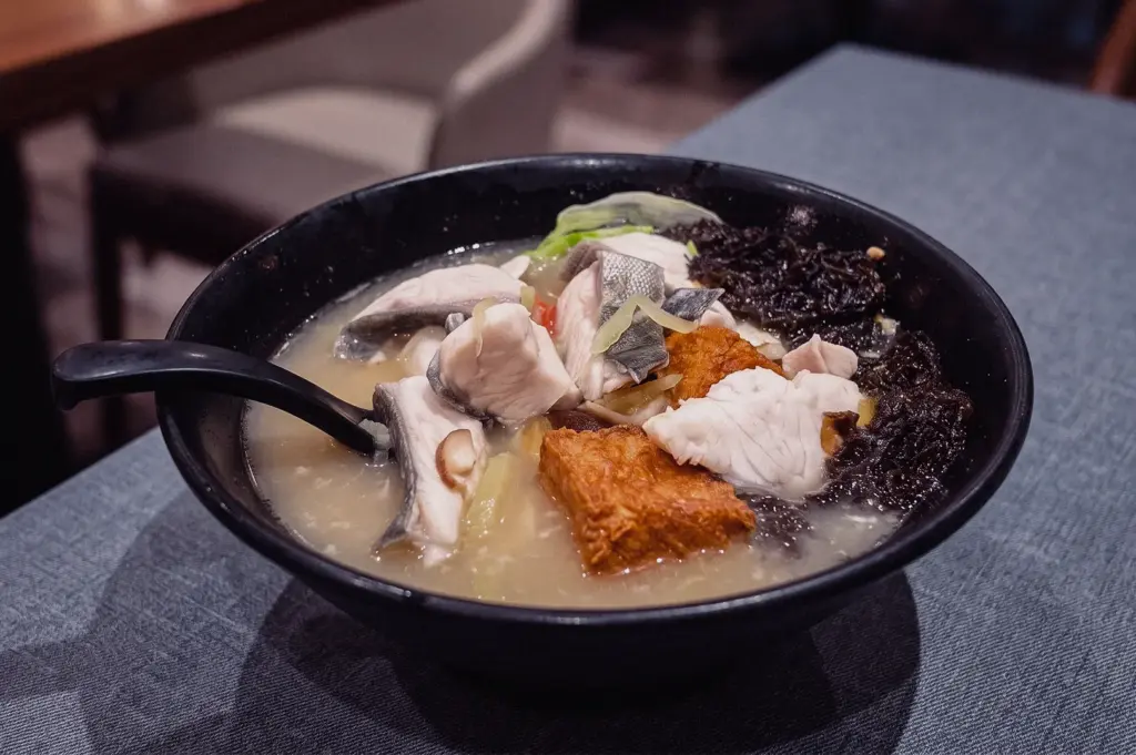 High-angle close-up shot of a black bowl filled with Grandpa's Pomfret Soup featuring tender pomfret slices, fried tofu, seaweed, and ginger in collagen broth.