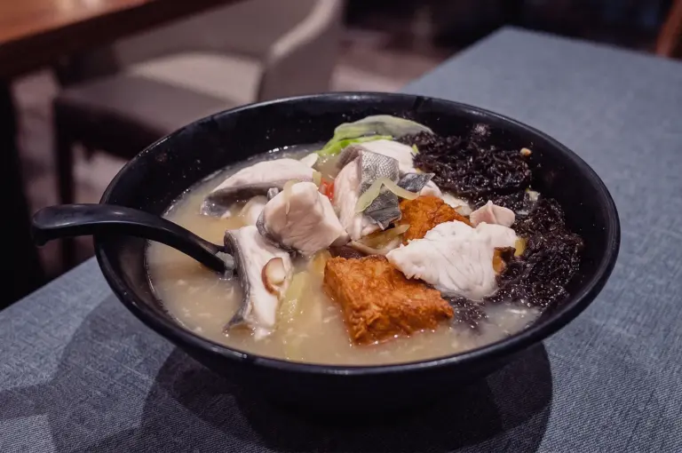 High-angle close-up shot of a black bowl filled with Grandpa's Pomfret Soup featuring tender pomfret slices, fried tofu, seaweed, and ginger in collagen broth.