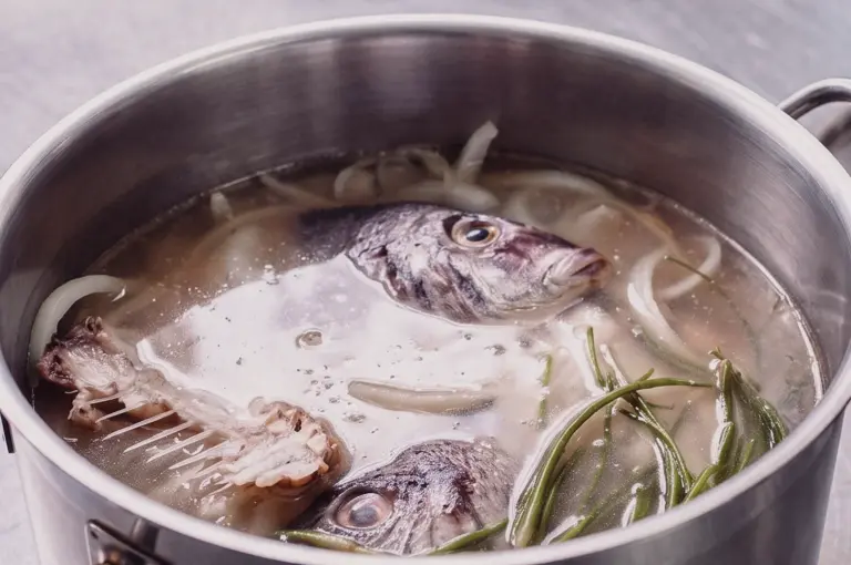 A high-angle close-up shot shows fish heads and clean fish bones simmering with herbs in a stainless steel stockpot to extract rich collagen for fish soup broth.