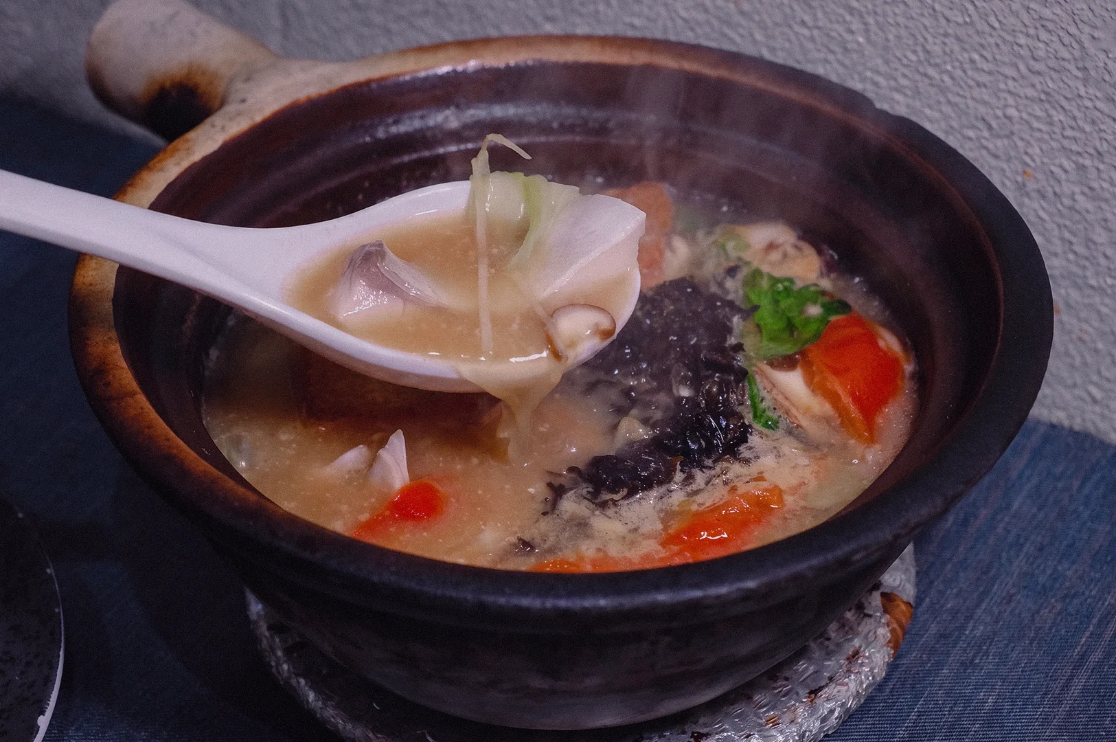 A high-angle close-up shot captures a spoon scooping tender pomfret fish slice and julienned ginger from a steaming claypot of rich collagen broth.