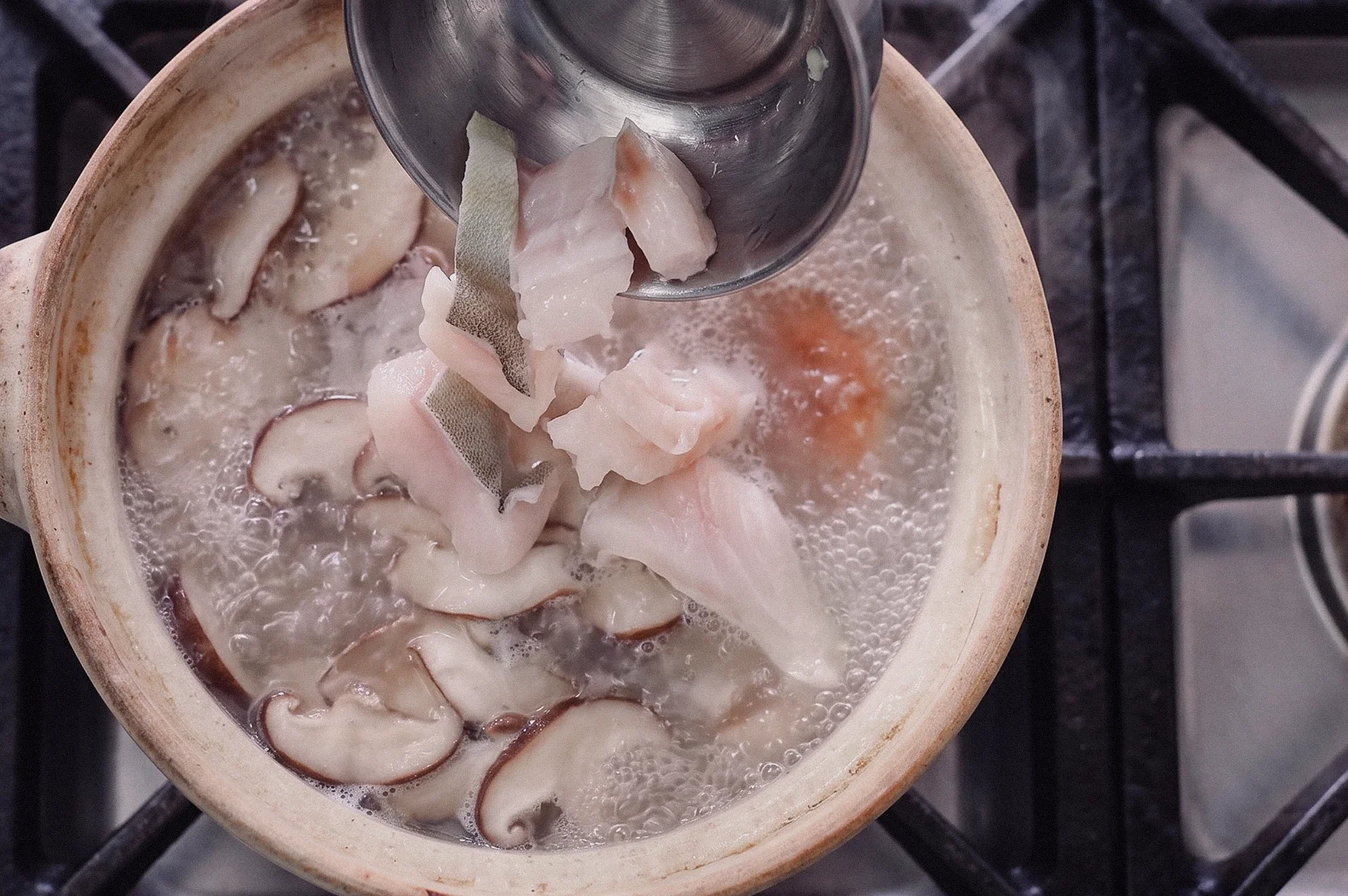A top-down overhead shot shows a metal ladle placing fresh pomfret fish slices into a claypot of simmering broth with sliced shiitake mushrooms.