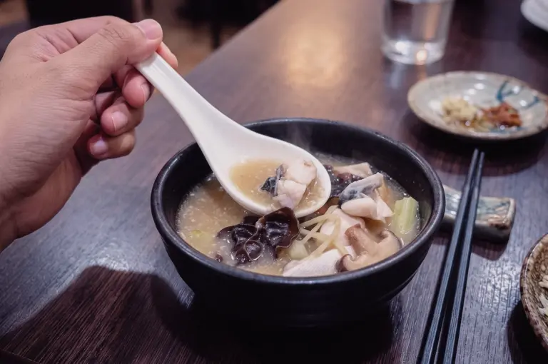 High-angle close-up shot capturing a hand using a white spoon to lift tender pomfret fish slices from a black bowl of steaming, freshly made Teochew fish soup.