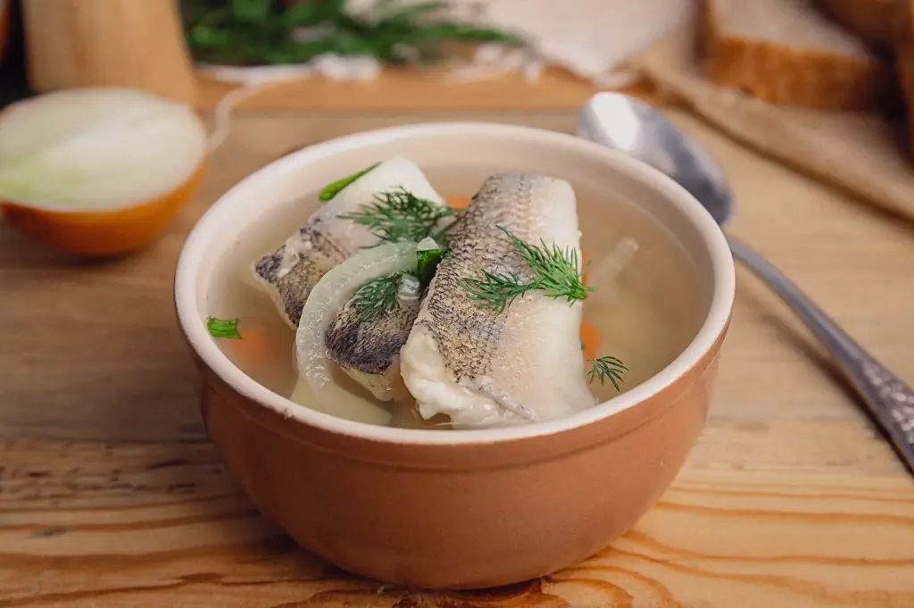 high-angle close-up shot shows a light, clear fish soup bowl garnished with fresh dill and tender fish fillets resting on a wooden tabletop.