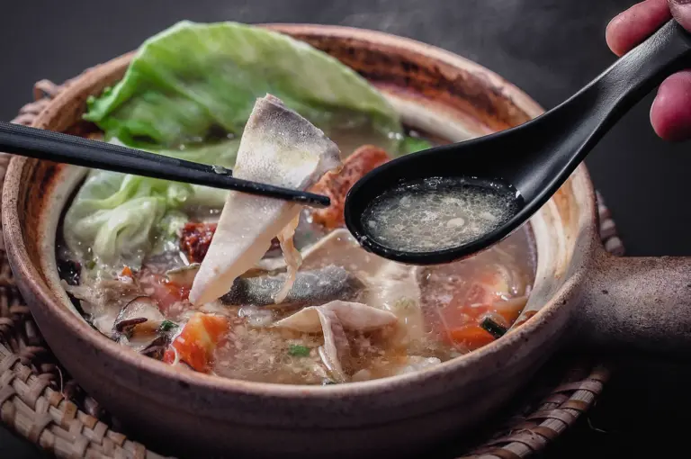 High-angle close-up shot capturing chopsticks lifting a tender pomfret fish slice over a hot claypot of rich broth alongside a spoon holding clear soup.