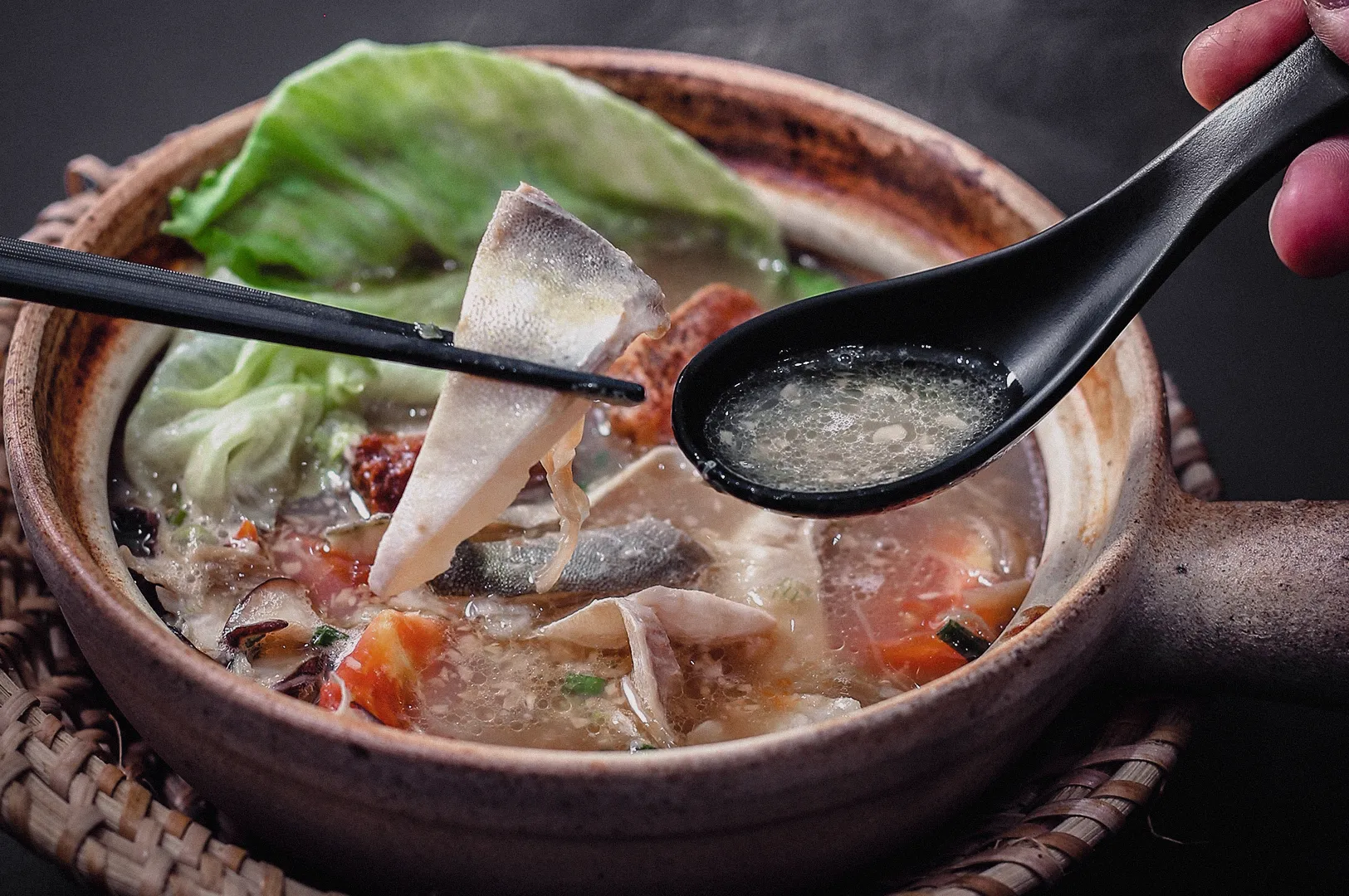 High-angle close-up shot capturing chopsticks lifting a tender pomfret fish slice over a hot claypot of rich broth alongside a spoon holding clear soup.