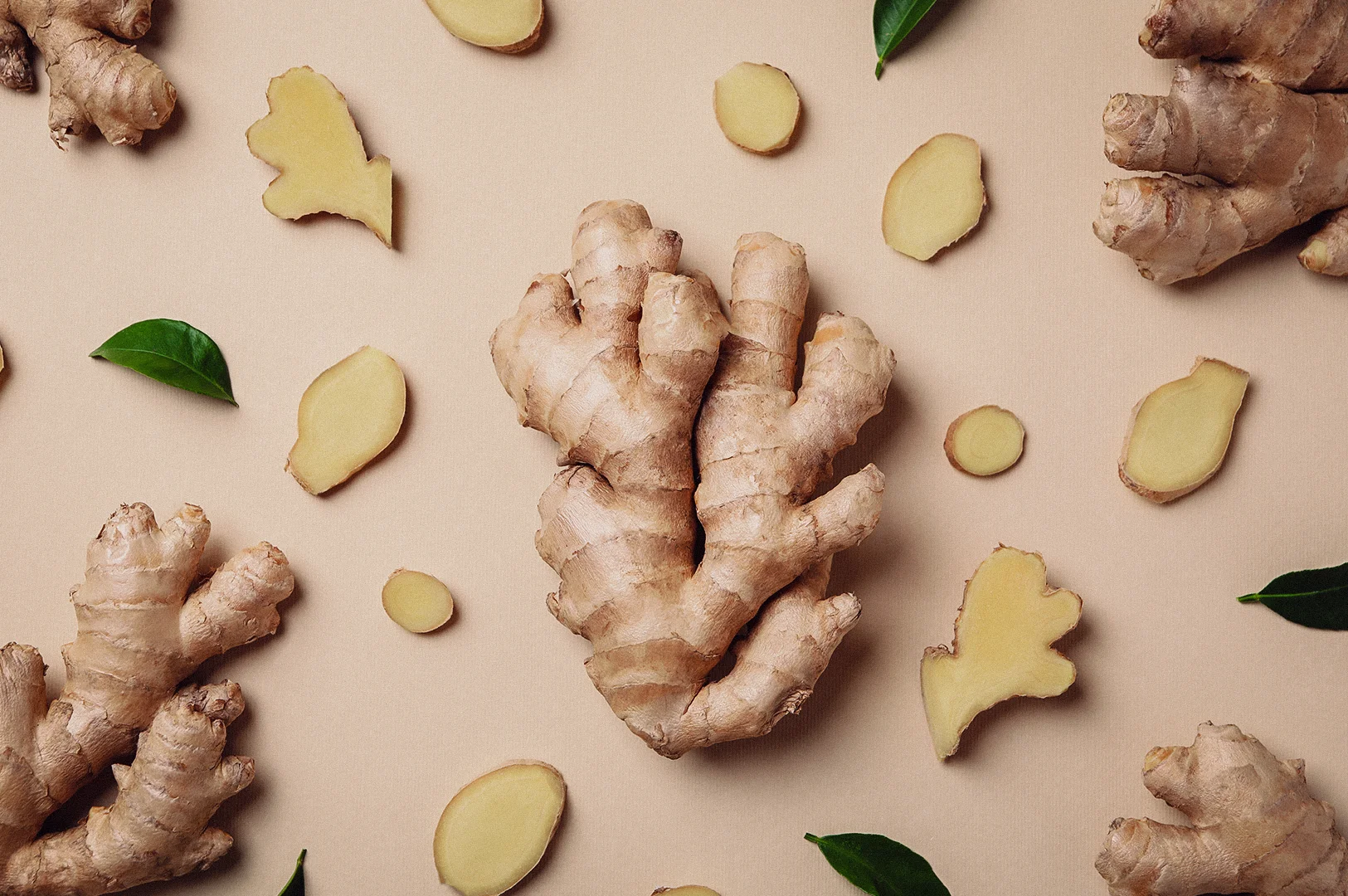 A top-down overhead flat lay shot displays whole fresh ginger roots and sliced ginger rounds arranged with green leaves on a light beige background.
