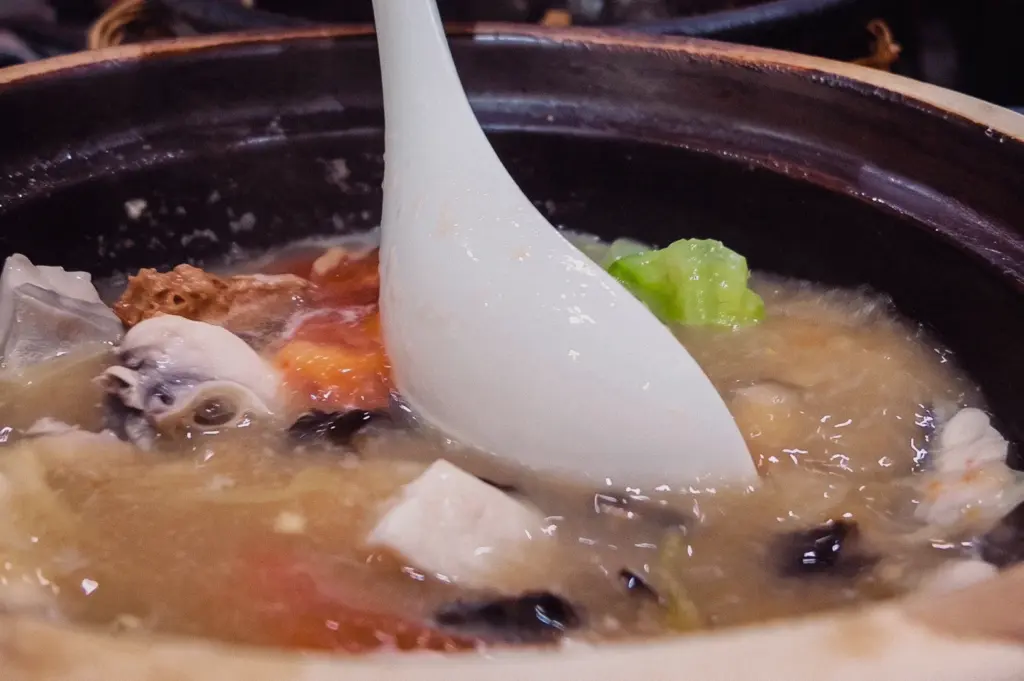 A close-up action shot captures a white spoon resting in a steaming claypot of collagen fish soup filled with tofu, cabbage, and mushrooms.