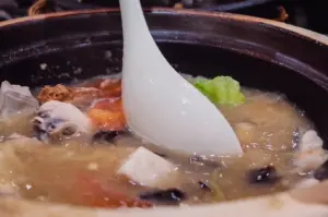 A close-up action shot captures a white spoon resting in a steaming claypot of collagen fish soup filled with tofu, cabbage, and mushrooms.