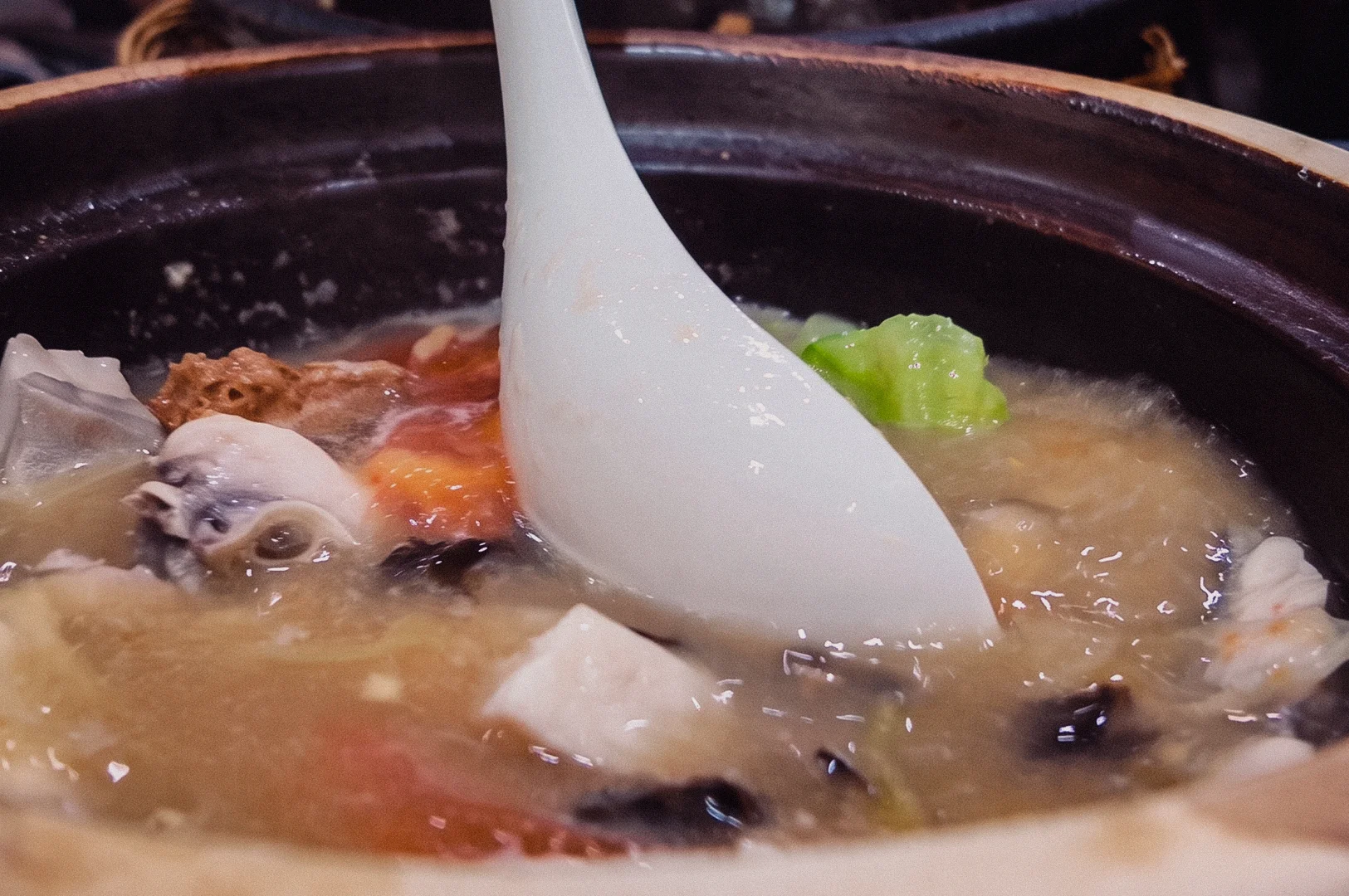 A close-up action shot captures a white spoon resting in a steaming claypot of collagen fish soup filled with tofu, cabbage, and mushrooms.