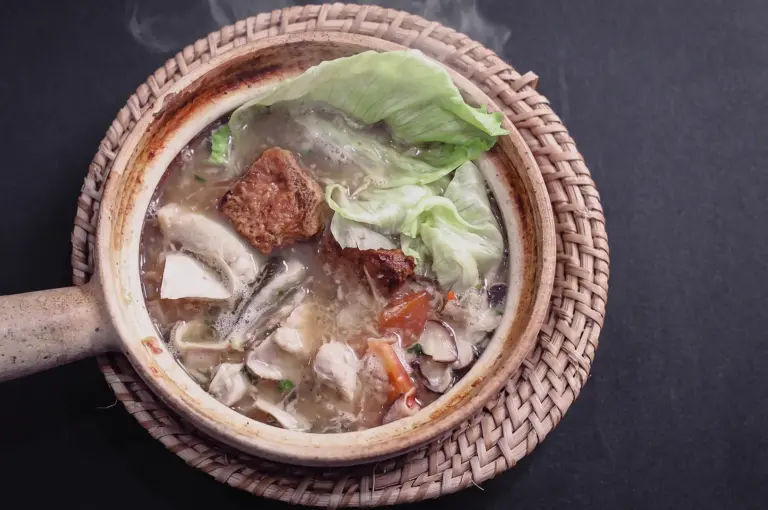 Top-down shot showcasing a piping hot claypot of pomfret fish soup with tofu puff, mushrooms, and lettuce resting on a woven rattan trivet.