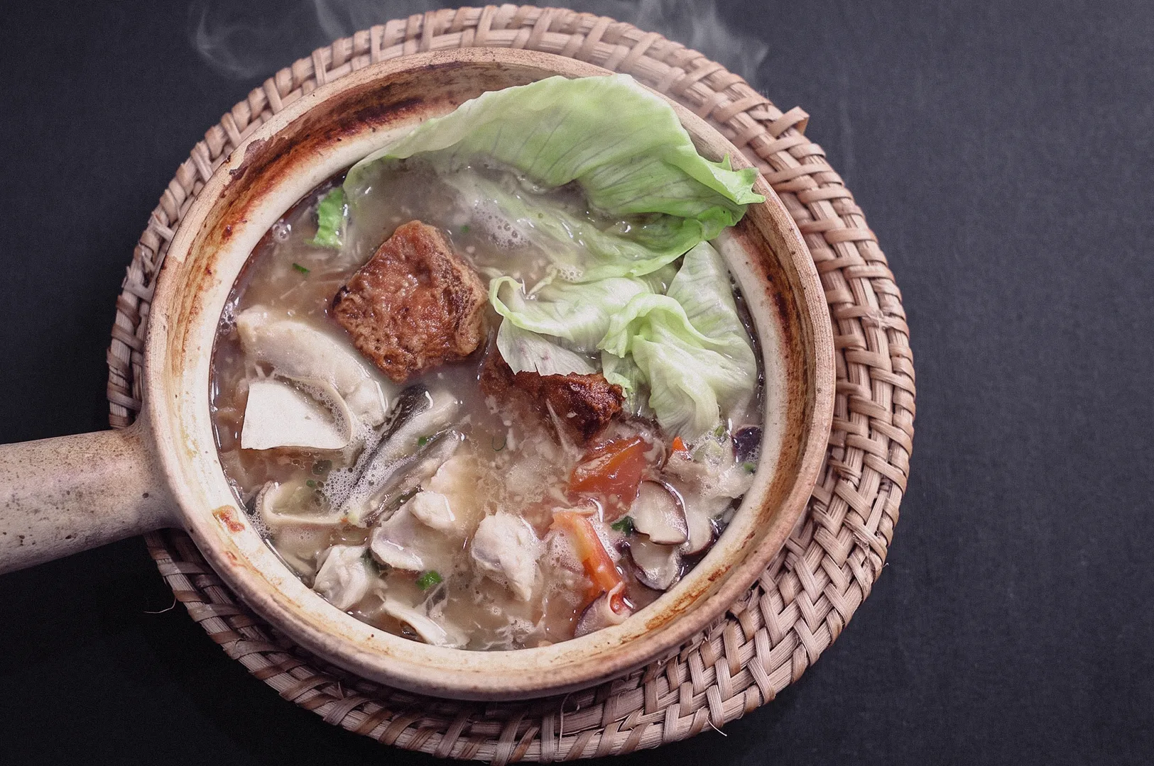 Top-down shot showcasing a piping hot claypot of pomfret fish soup with tofu puff, mushrooms, and lettuce resting on a woven rattan trivet.