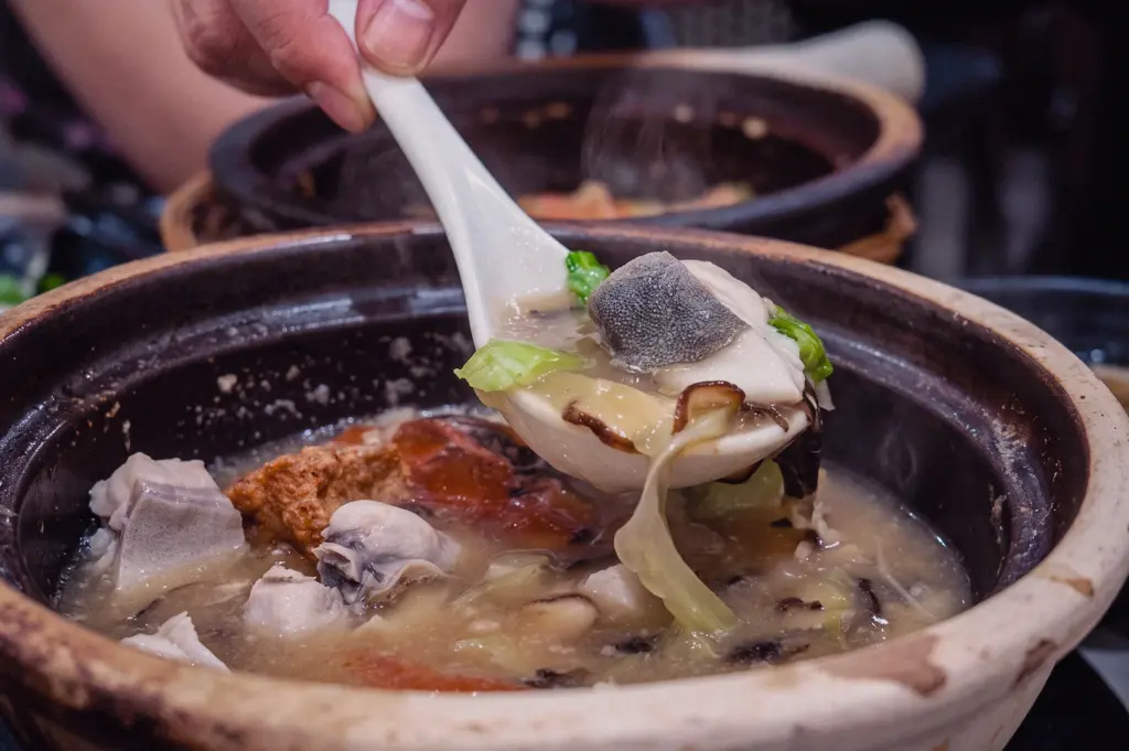 A close-up action shot captures a spoon scooping hot pomfret fish soup with tender fish slices and fresh greens from a traditional claypot.
