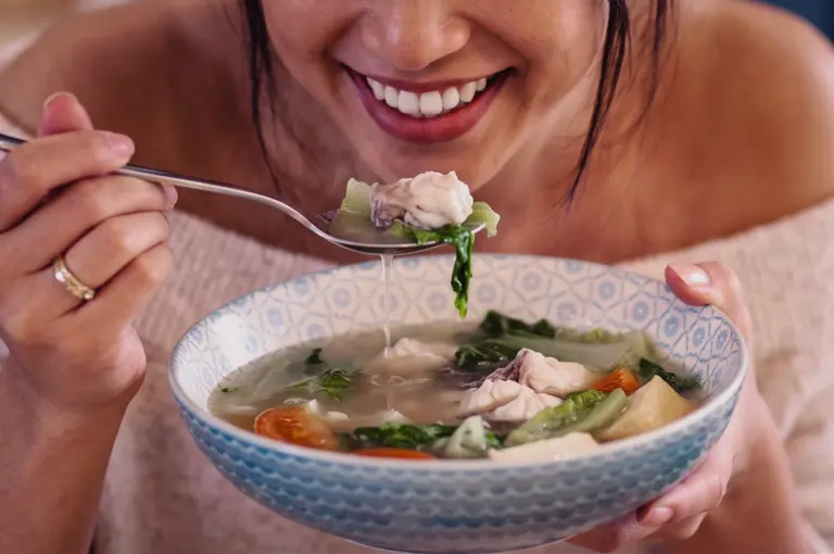 A close-up medium shot captures a smiling woman eating a spoon of clean, light fish soup with tender fish slices and fresh leafy greens.