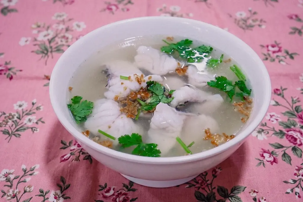 Three-quarter view of a white ceramic bowl filled with delicate clear fish soup, tender white fish slices, and fresh cilantro leaves on a floral table cloth.