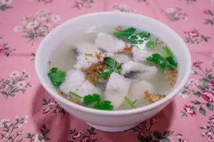 Three-quarter view of a white ceramic bowl filled with delicate clear fish soup, tender white fish slices, and fresh cilantro leaves on a floral table cloth.