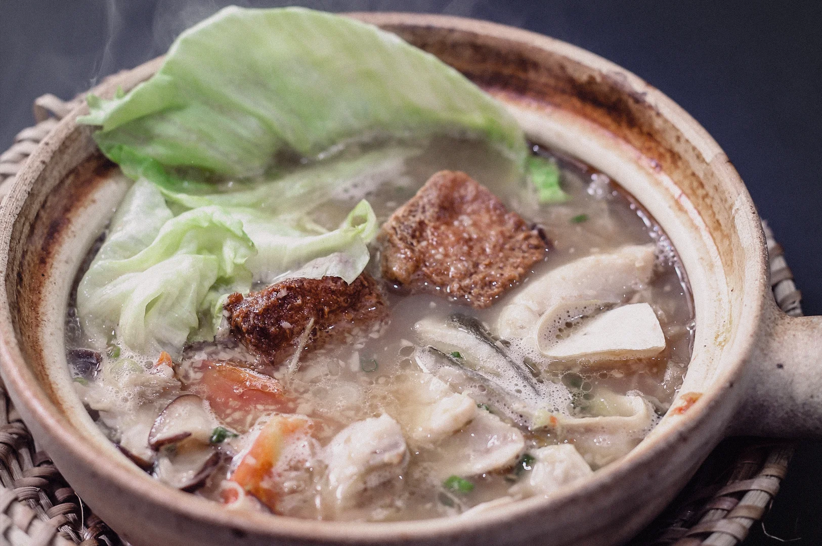 A high-angle close-up shot highlights tender pomfret fish slices, leafy greens, and fried tofu simmered in a steaming claypot of collagen fish broth.