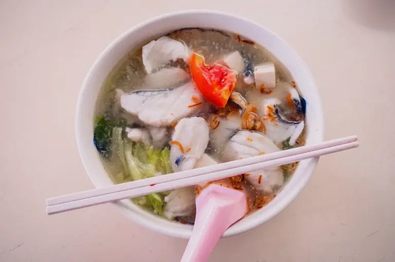 A top-down overhead flat lay shot displays a bowl of clear fish soup served with chopsticks, pink spoon, sliced fish, tomatoes, and greens on a dining table.