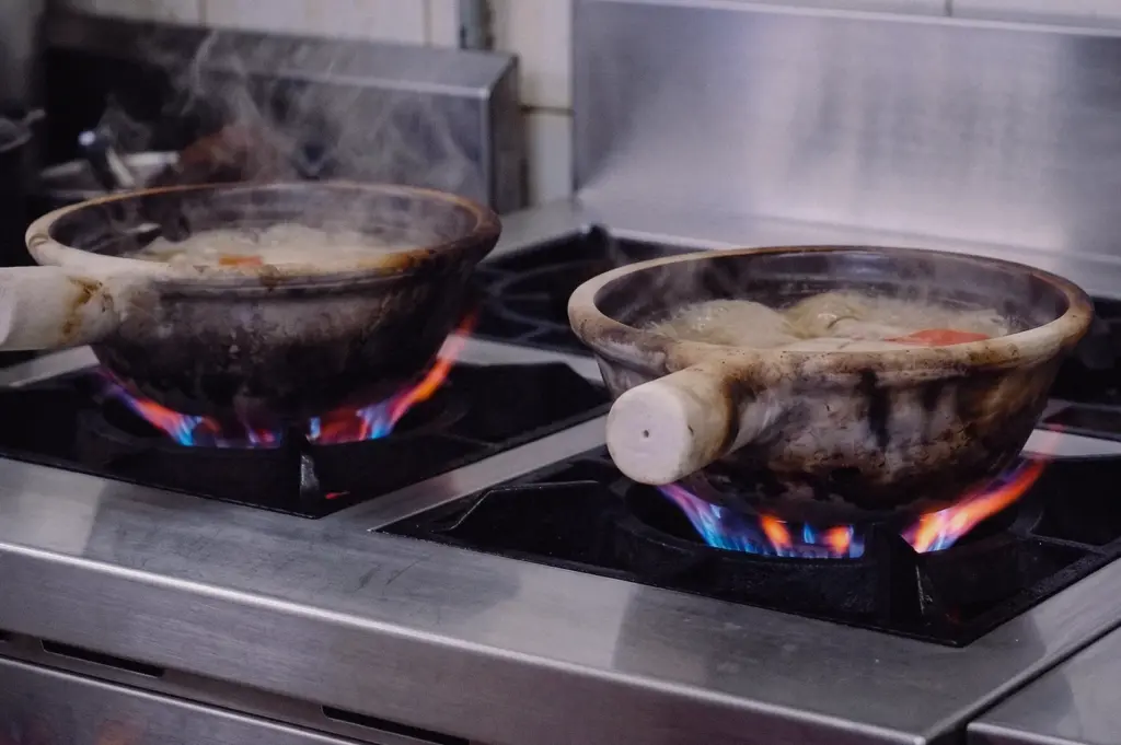 A low-angle three-quarter shot captures two claypots simmering over vibrant gas stove flames in a commercial kitchen.