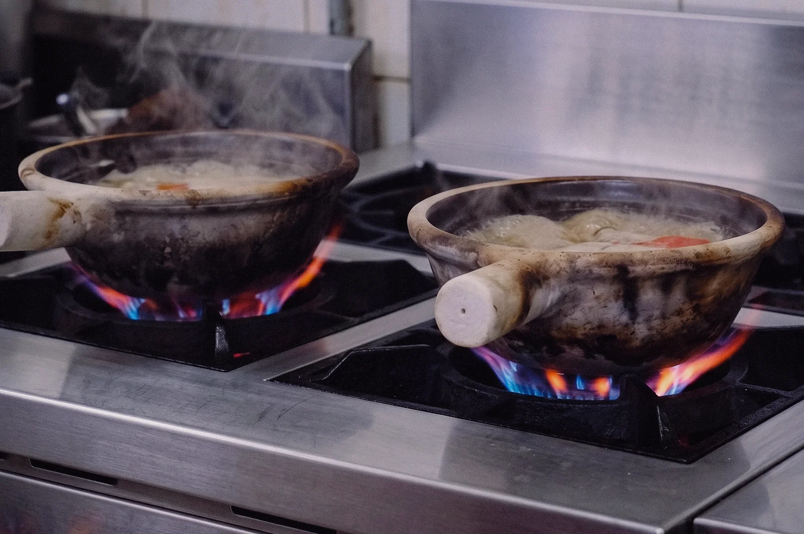 A low-angle three-quarter shot captures two claypots simmering over vibrant gas stove flames in a commercial kitchen.