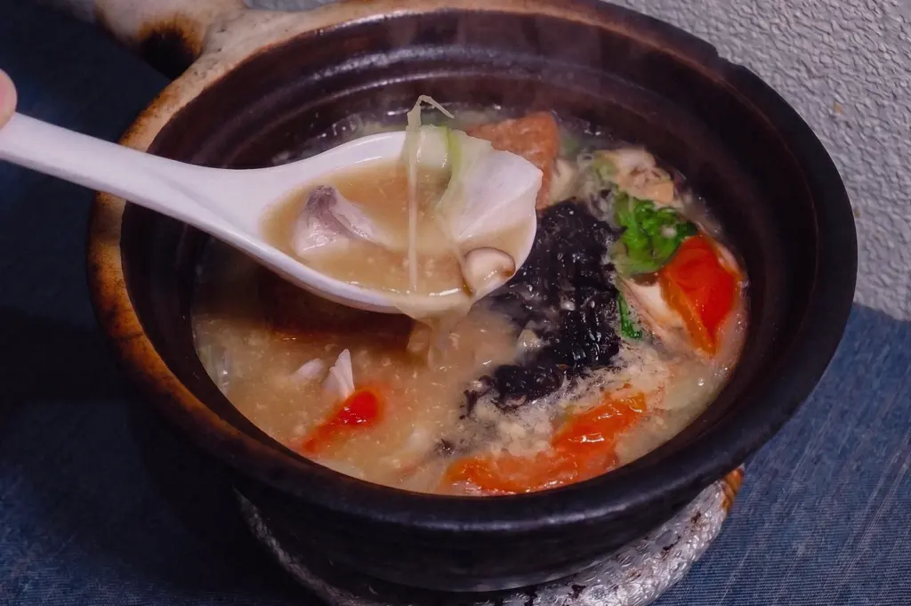 High-angle close-up shot capturing a white ceramic spoon scooping tender pomfret fish slices from a bubbling claypot of rich, collagen-infused Teochew broth.