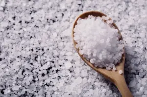 A high-angle close-up shot shows coarse sea salt crystals heaped onto a wooden spoon over a scattered bed of salt grains.
