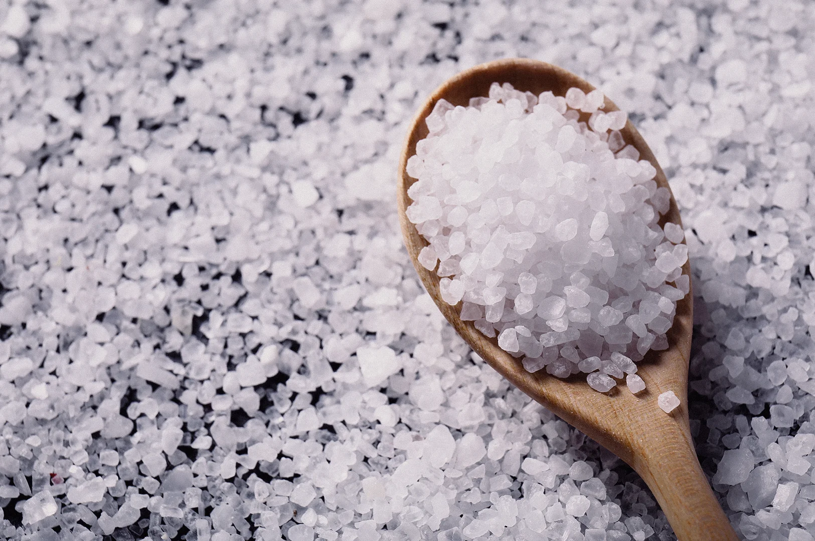 A high-angle close-up shot shows coarse sea salt crystals heaped onto a wooden spoon over a scattered bed of salt grains.