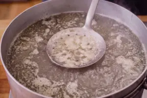 A high-angle close-up shot shows a large perforated skimmer spoon lifting grey scum and impurities from a pot of simmering fish broth.