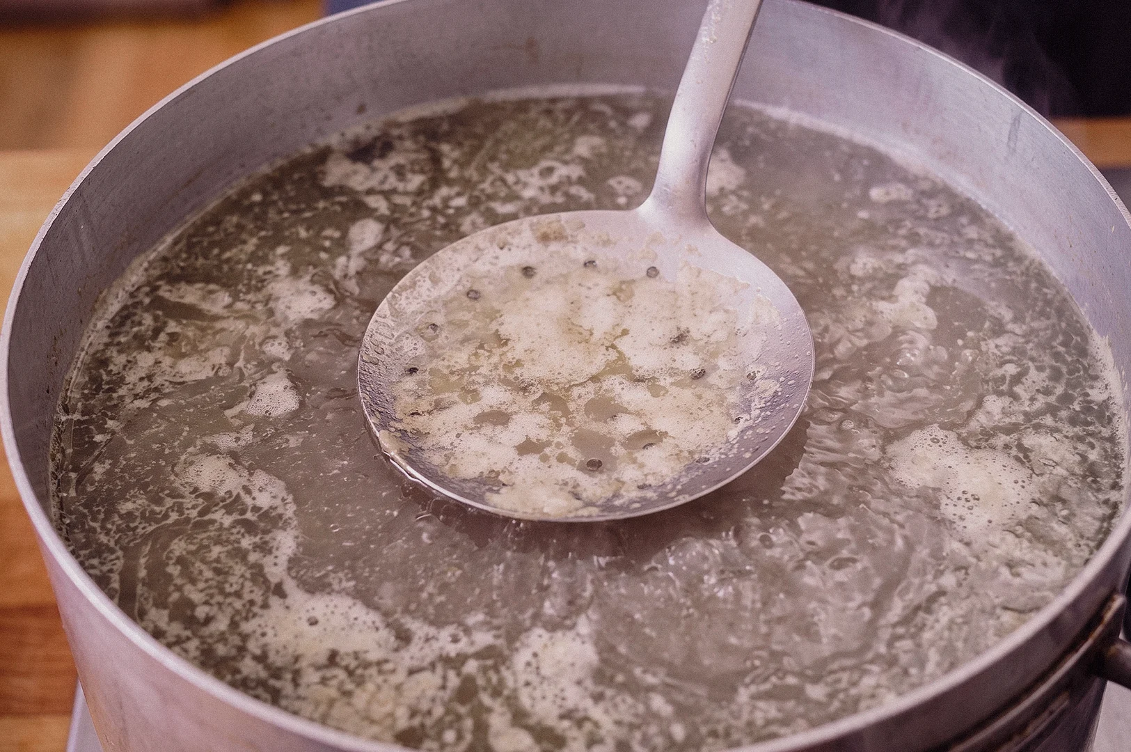 A high-angle close-up shot shows a large perforated skimmer spoon lifting grey scum and impurities from a pot of simmering fish broth.