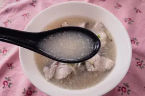 A high-angle close-up shot captures a black spoon scooping a sample of cloudy, opaque fish soup broth above a bowl filled with fish slices set against a floral pink tablecloth.