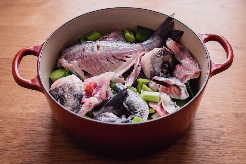 High-angle three-quarters shot looking into a red cast-iron pot filled with fresh raw fish frames, fish heads, and chopped scallions ready to be simmered into broth.