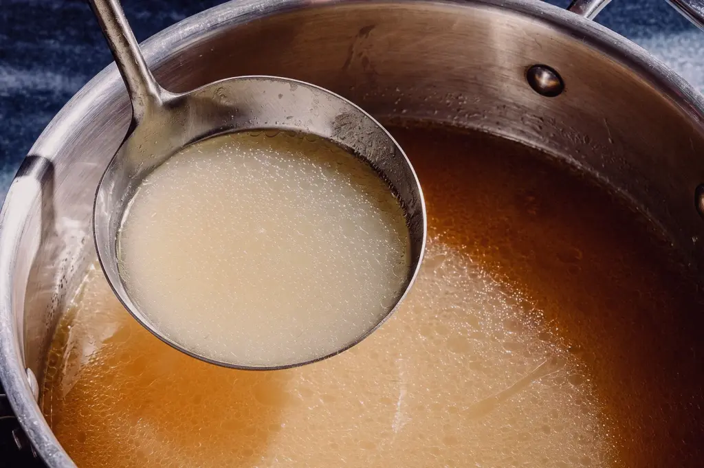 A high-angle close-up shot shows a metal ladle lifting clear, unseasoned fish stock from a stainless steel pot